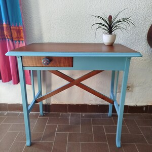Puede incluir: Una mesa de madera azul claro con un solo cajón y una estructura de soporte en forma de cruz. La mesa tiene una tapa marrón y una pequeña planta en una maceta blanca está sentada sobre la mesa.