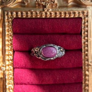 May include: A silver ring with a pink oval gemstone set in a textured band. The ring is displayed in a red velvet ring box.