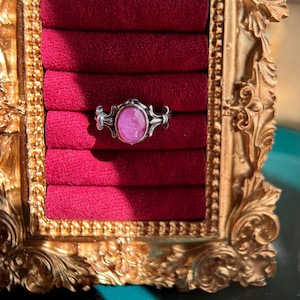 May include: A silver ring with a pink oval gemstone set in a decorative filigree design. The ring is displayed in a velvet-lined ring box.