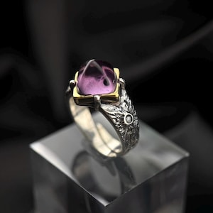 May include: A silver ring with a purple gemstone set in a gold frame. The ring band is decorated with a textured design.
