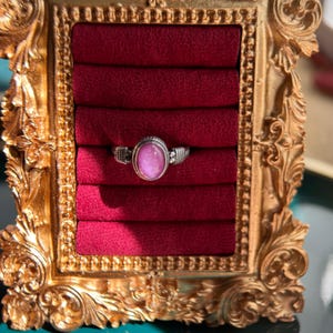 Puede incluir: Un anillo de plata con una piedra ovalada rosa engastada en una banda de plata decorativa. El anillo se muestra en una caja de anillo de terciopelo rojo.