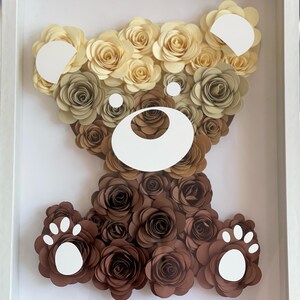May include: A framed paper flower art piece depicting a brown bear. The bear is made of brown and cream paper flowers. The bear has a white nose and white paw prints.
