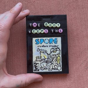 Puede incluir: Un objeto rectangular negro con el texto "THE BODY KEEPS THE" en letras de cuentas. Debajo, una colorida ilustración de criaturas con la palabra "SPORE" y "creador de criaturas" se muestra. El objeto es sostenido por una mano.