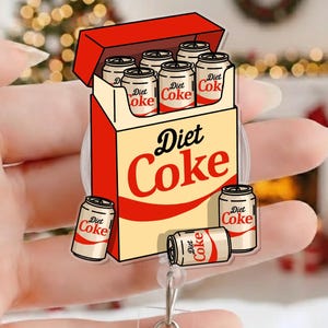 May include: A Diet Coke badge reel showing a box of Diet Coke cans. The box is red and beige with the words "Diet Coke" in red and black. Several Diet Coke cans are scattered around the box.