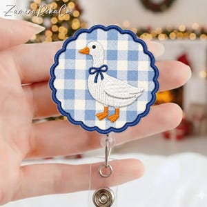 May include: A circular badge reel featuring an embroidered white goose with an orange beak and feet, wearing a dark blue bow. The background is a light blue and white gingham check pattern with a scalloped dark blue embroidered border edge.