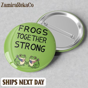 May include: A green button with the text "FROGS TOGETHER STRONG" and two cartoon frogs wearing bandanas with an American flag design. The button has a silver pin back.