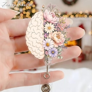 Könnte beinhalten: Ein Ausweis-Rollhalter mit einem Gehirndesign und floralen Akzenten. Das Gehirn ist beige, und die Blumen sind rosa, lila und weiß. Die Rolle hat einen transparenten Kunststoffriemen und einen Metallclip.