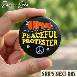 May include: A black button with the words "UNPAID PEACEFUL PROTESTER" in yellow and orange, with a peace symbol. White daisy accents surround the text. The button is held in a hand.