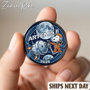 May include: A round, blue and silver Artemis II commemorative coin featuring Snoopy in an orange spacesuit, a spacecraft, and the moon. The coin has the text "ARTEMIS II" and "2026".