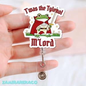 May include: A badge reel featuring a cartoon frog dressed as a knight, holding a sword. The frog wears a red cape and a white outfit. Text on the badge reads "T'was the Tylenol m'Lord". The reel has a clear plastic strap and a metal button.