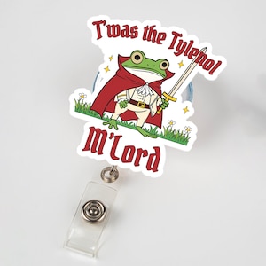 May include: A badge reel with a cartoon frog wearing a red cape and holding a sword. The text on the badge reel reads "T'was the Tylenol m'lord". The frog is standing on green grass with small white flowers.