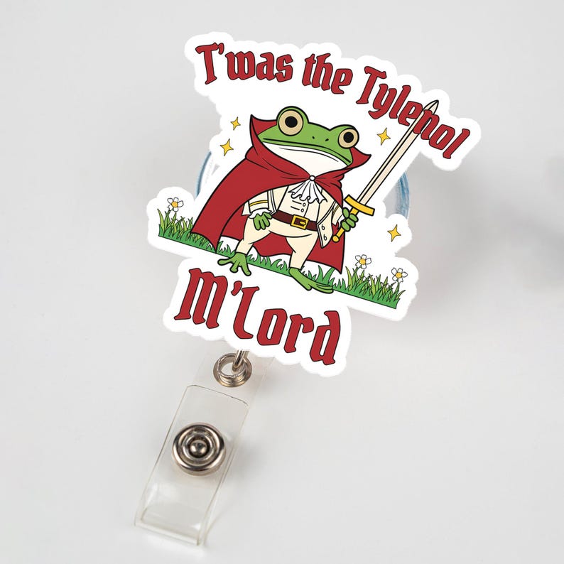 May include: A badge reel with a cartoon frog wearing a red cape and holding a sword. The text on the badge reel reads "T'was the Tylenol m'lord". The frog is standing on green grass with small white flowers.
