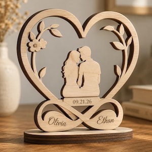 May include: A wooden heart-shaped decorative piece featuring a silhouette of a couple kissing, with floral and leaf accents. The design includes the date "09.21.26" and the names "Olivia" and "Ethan" on an infinity symbol base.