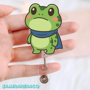 May include: A green frog cartoon character badge reel with a blue cape and a sad expression. The badge reel has a clear plastic strap and a metal clip. The text "ZAMIRAREKAKO" is at the bottom.