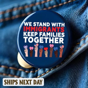 May include: A round, navy blue button with the text "WE STAND WITH IMMIGRANTS KEEP FAMILIES TOGETHER" in white and red. The button features illustrations of hands holding hearts and peace signs. The button is set against a denim background.