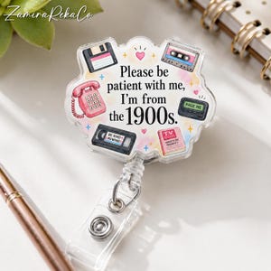 May include: A clear acrylic badge reel featuring a scalloped edge and colorful illustrations of a floppy disk, cassette tape, rotary phone, pager, VHS tape, and TV Guide. The text reads: Please be patient with me, I'm from the 1900s.