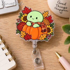 May include: A clear acrylic badge reel featuring a cartoon light green creature wearing a plaid scarf and sitting inside a bright orange pumpkin. The design is surrounded by colorful autumn leaves, yellow sunflowers, and red berries.
