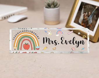 May include: A rectangular desk nameplate with the text "Mrs. Evelyn" in black. The nameplate features a rainbow, apple, and pencil design with the words "Teach Love Inspire". The background is beige with small colourful illustrations.