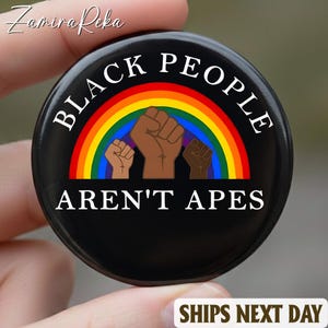 Può includere: Un bottone nero con la scritta bianca "BLACK PEOPLE AREN'T APES". Il bottone presenta un design arcobaleno e tre pugni alzati in diverse tonalità di marrone. Il bottone è tenuto in mano.