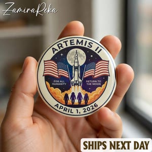 May include: A round button featuring the Artemis II mission. The design includes a rocket, American flags, and the text "FOR ALL HUMANITY" and "RETURN TO THE MOON." The date "APRIL 1, 2026" is also displayed. The button is held in a hand.