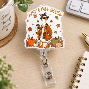 May include: A clear acrylic badge reel featuring a printed illustration of a goose wearing a leopard print headband, sunglasses, and a plaid scarf while holding a coffee cup. The text reads JUST A FALL GOOSE, surrounded by pumpkins and autumn leaves.