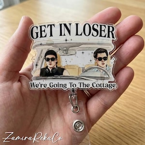 May include: A clear badge reel featuring the text "GET IN LOSER" above an illustration of two people in a car. The text "We're Going To The Cottage" is below the car illustration. The badge reel is held in a hand.