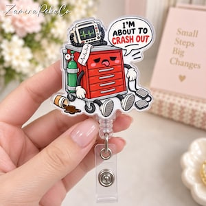 May include: An acrylic badge reel featuring a cartoon red medical crash cart with a green oxygen tank, a heart monitor, and a spilled coffee cup. The cart has a tired face and a speech bubble with the text I'M ABOUT TO CRASH OUT in black and red.