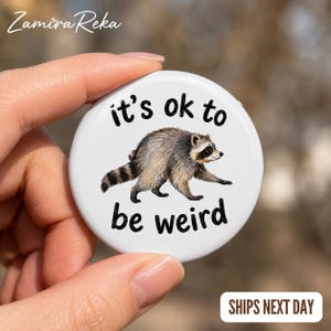 Op de afbeelding: Een ronde witte button met een gedetailleerde illustratie van een wasbeer in het midden. De tekst "it's ok to be weird" is in zwarte, afgeronde letters boven en onder de wasbeerafbeelding op het gladde oppervlak gedrukt.