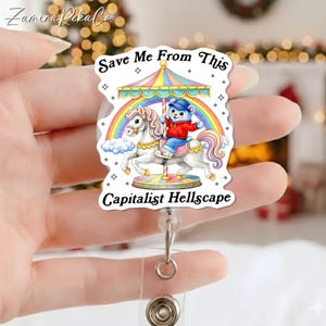 May include: A clear, rectangular badge reel with a carousel design. The carousel features a white horse, a bear in a red jacket, and a rainbow. Text on the badge reel reads "Save Me From This Capitalist Hellscape."