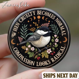 Puede incluir: Un botón redondo negro con el texto "WHEN CRUELTY BECOMES NORMAL COMPASSION LOOKS RADICAL" que rodea una ilustración detallada de un pájaro posado en una rama con flores. El botón tiene un borde decorativo con lunas y estrellas.
