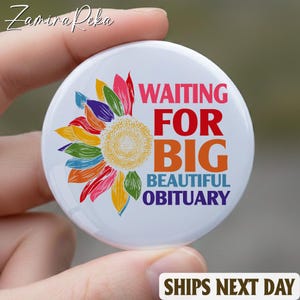 以下が含まれることがあります： カラフルなヒマワリのデザインと「WAITING FOR BIG BEAUTIFUL OBITUARY」という文字が赤、オレンジ、紫で書かれた白いボタン。ボタンは手で持たれています。