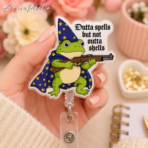 Può includere: Un porta badge in acrilico raffigurante una rana verde con un cappello da mago blu e un mantello con stelle gialle. La rana tiene in mano un fucile marrone. Il testo "Dutta spells but not outta shells" è scritto in un carattere gotico nero.