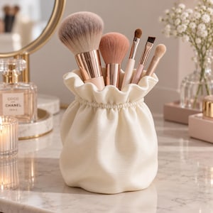 Peut inclure: Un porte-pinceaux de maquillage en céramique crème texturée, en forme de bourse froncée. Le récipient contient cinq pinceaux de maquillage variés avec des viroles en or rose et des poils doux dans des tons de brun, beige et rose.