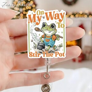 May include: A badge reel with a cartoon frog character holding a wooden spoon and stirring a pot. The text on the badge reads "On My Way To Stir The Pot" in orange and white lettering. The frog is wearing overalls and a shirt.