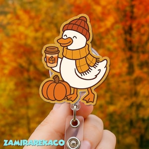 May include: A fall-themed badge reel featuring a cartoon duck wearing a knit cap and scarf, holding a coffee cup and standing next to a pumpkin. The duck is white with orange accents. The background is a blurred autumn scene.