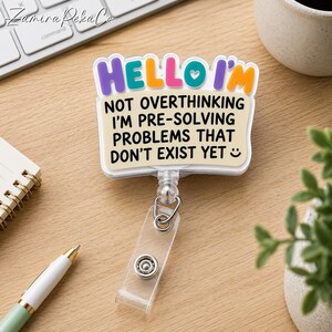 Op de afbeelding: Een rechthoekige badgehouder met een crèmekleurige achtergrond en zwarte tekst: HELLO I'M NOT OVERTHINKING I'M PRE-SOLVING PROBLEMS THAT DON'T EXIST YET. De bovenkant heeft kleurrijke, verhoogde letters in paars, turquoise, roze en oranje.