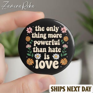 May include: A black circular button featuring the words "the only thing more powerful than hate is love" in white, with floral designs in pink, orange, and white. The button is held by a hand.