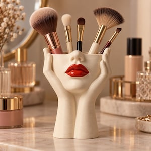 May include: A cream-coloured makeup brush holder shaped like a face with hands cupping the chin. The holder features prominent red lips and contains five various makeup brushes with metallic handles, including a large powder brush and a fan brush.