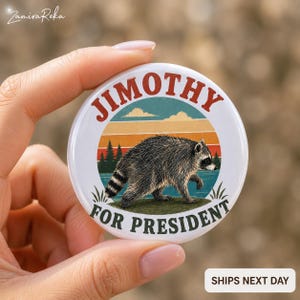 Op de afbeelding: Een ronde witte button met een afbeelding van een wasbeer die door een landschap loopt met een retro zonsondergang, dennenbomen en water. De tekst JIMOTHY staat in een boog bovenaan en FOR PRESIDENT onderaan in donkere letters.