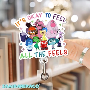 Puede incluir: Un portatarjetas colorido con personajes de la película "Del revés". El texto dice "It's okay to feel all the feels". Los personajes representan diferentes emociones. El portatarjetas tiene un soporte de plástico transparente y un clip metálico.