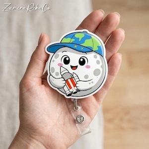 Puede incluir: Un carrete de insignia blanco con un personaje de dibujos animados de la luna que lleva una gorra azul y verde con el tema de la Tierra. La luna sostiene un cohete y tiene una cara sonriente con mejillas rosadas. El carrete de la insignia tiene una correa de plástico transparente y un botón plateado.