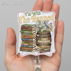 Puede incluir: Un portatarjetas rectangular con el texto "I'm With The Banned". El diseño incluye una pila de libros, un barril de madera lleno de flores y el texto "I Read Banned Books".