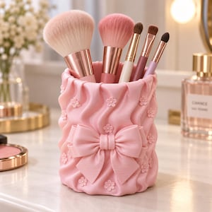 May include: A cylindrical light pink makeup brush holder featuring a textured, ruffled surface adorned with small raised flower accents and a prominent central bow. Five makeup brushes with metallic handles are placed inside the container.