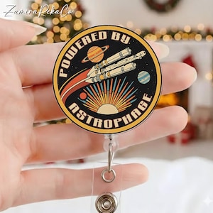 Könnte beinhalten: Ein runder Ausweis-Rollhalter mit Retro-Weltraum-Thema. Das Design zeigt ein Raumschiff, Planeten und die Worte "POWERED BY ASTROPHAGE" in kreisförmiger Anordnung. Die Rolle hat einen transparenten Kunststoffriemen und einen Metallclip.