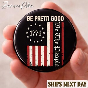 May include: A black round button with a distressed American flag design. The flag features red and white stripes, a circle of stars with the year 1776, and the text "BE PRETTI GOOD" and "We The People".