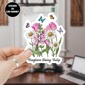 May include: A sticker or car magnet featuring a floral design with pink tulips, white daisies, and purple foxgloves, along with several butterflies. The text "Foxglove Daisy Tulip" is printed at the bottom. The sticker is held by a hand.