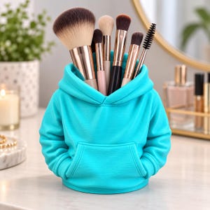 Peut inclure: Un organisateur de bureau turquoise vif en forme de sweat-shirt à capuche miniature, doté d'une capuche, de manches longues et d'une poche kangourou. Il contient divers pinceaux de maquillage avec des viroles en or rose.
