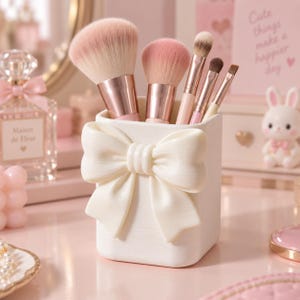 Puede incluir: Un porta brochas de maquillaje cuadrado de color blanco con un gran lazo esculpido en la parte frontal. El recipiente contiene cinco brochas con virolas de oro rosa y cerdas suaves con puntas rosadas. Tiene un acabado mate liso y bordes redondeados.