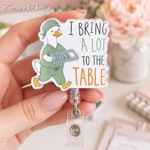 May include: A clear acrylic badge reel featuring a cartoon duck dressed in green surgical scrubs and a matching cap. The duck holds a tray of medical instruments. The text reads I BRING A LOT TO THE TABLE in black and orange lettering.