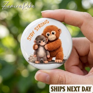 May include: A round white button featuring two cartoon monkeys hugging. The text "STAY STRONG" is in orange. The button is held in a hand against a blurred green background. The words "SHIPS NEXT DAY" are at the bottom.